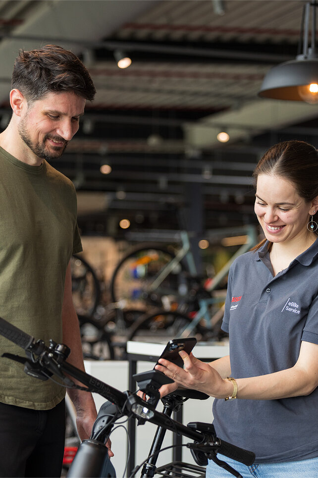 Zwei Personen stehen in einem Fahrradgeschäft. Eine Person trägt ein olivgrünes T-Shirt und schwarze Hosen, die andere Person trägt ein graues Poloshirt mit Bosch-Logo und hält ein Smartphone. Im Hintergrund sind mehrere Fahrräder und eine moderne Einrichtung zu sehen.