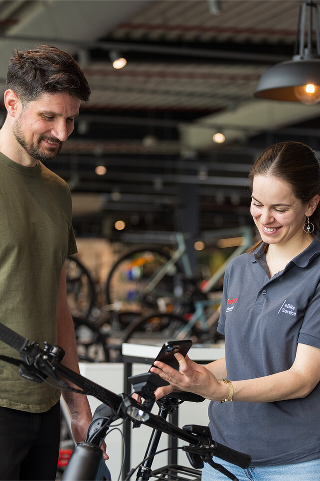 Zwei Personen stehen in einem Fahrradgeschäft. Eine Person trägt ein olivgrünes T-Shirt und schwarze Hosen, die andere Person trägt ein graues Poloshirt mit Bosch-Logo und hält ein Smartphone. Im Hintergrund sind mehrere Fahrräder und eine moderne Einrichtung zu sehen.