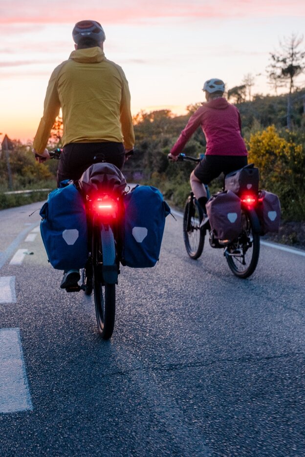 Zwei Personen fahren mit E-Bikes auf einer Straße bei Sonnenuntergang. Beide E-Bikes sind mit Gepäcktaschen ausgestattet und haben rote Rücklichter. Die Person links trägt eine gelbe Jacke und die Person rechts eine rote Jacke. Die Umgebung ist ländlich mit Bäumen und Sträuchern.