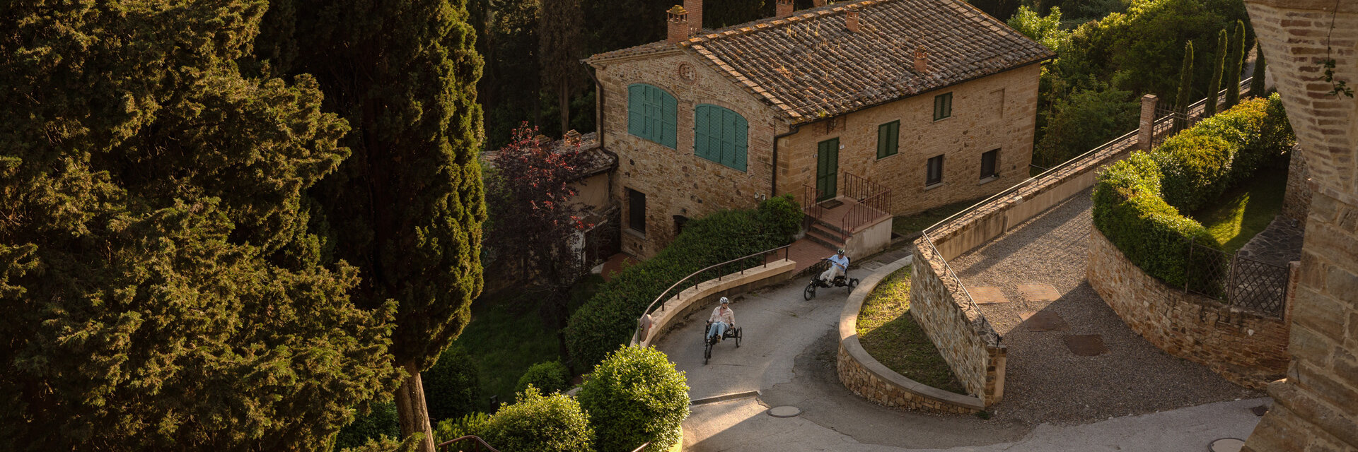 Due persone viaggiano in eBike lungo una strada tortuosa, passando davanti a una casa di pietra immersa in un paesaggio collinare con cipressi.