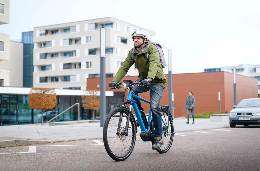 Een eBike-rijder in een groene jas rijdt op een blauw eBike door een straat, op de achtergrond zijn gebouwen, een voetganger en een geparkeerde auto te zien.