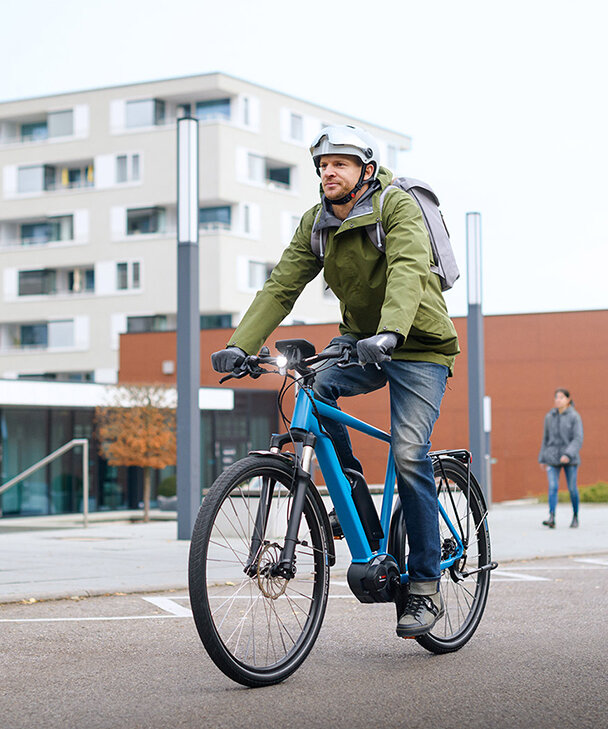 Een eBike-rijder in een groene jas rijdt op een blauw eBike door een straat, op de achtergrond zijn gebouwen, een voetganger en een geparkeerde auto te zien.