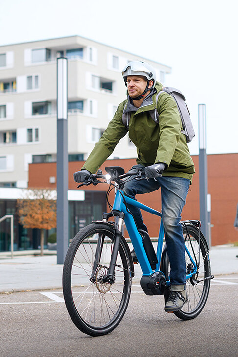 Un eBike piloto con chaqueta verde circula en un eBike azul por una carretera, al fondo se distinguen edificios, un peatón y un Auto estacionado.