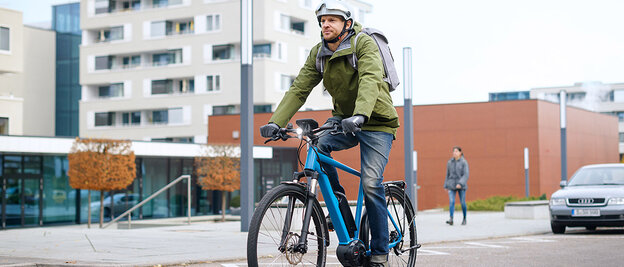 Un utilisateur d'[eBike] conduit dans une rue en veste verte sur un [eBike] bleu, en arrière-plan, vous pouvez voir des bâtiments, un piéton et une voiture garée.
