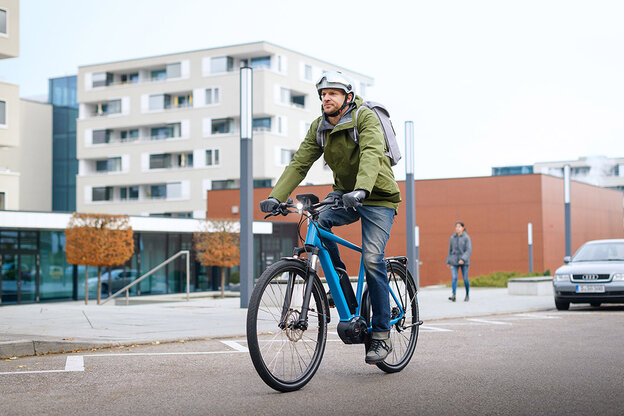 En eBike-cyklist i en grön jacka kör längs en gata på en blå eBike. I bakgrunden syns byggnader, en fotgängare och en parkerad bil.