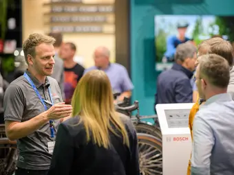 A group of people gathers at a Bosch booth at a trade fair. A Bosch employee is explaining something to them, with the handlebars of a racing bike visible in the background.