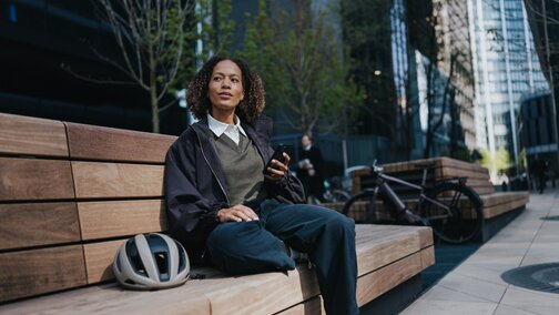 Une femme est assise sur un banc, un téléphone portable à la main. À côté d’elle se trouve un casque de vélo blanc, tandis qu’en arrière-plan, un VAE moderne avec un moteur de moyeu Bosch est visible.