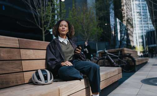 A woman sits on a bench with her mobile phone in hand. Next to her is a white bicycle helmet, and in the background, you can see a modern eBike with a hub motor from Bosch.