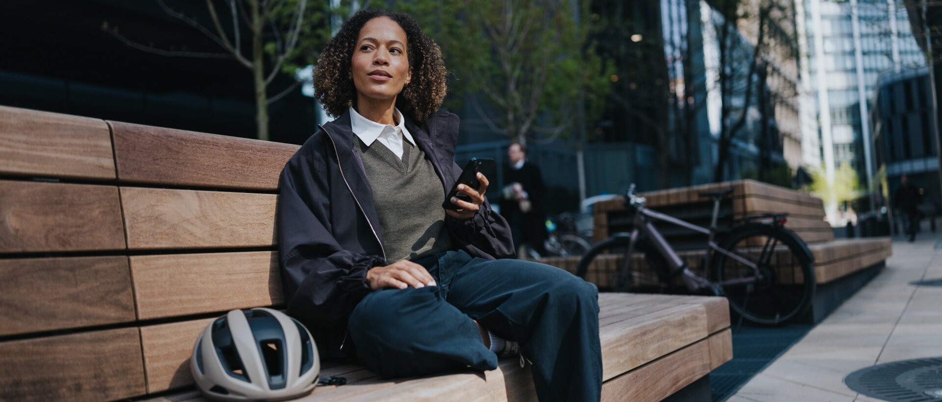 A woman sits on a bench with her mobile phone in hand. Next to her is a white bicycle helmet, and in the background, you can see a modern eBike with a hub motor from Bosch.